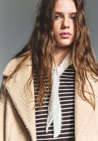 Beige ullkappa med mjuk textur, bärs över en bruna och vita randiga topp. En ljus scarf är knuten runt halsen.