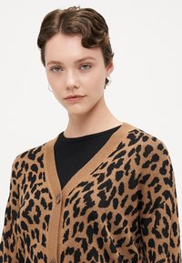 Knit cardigan in tan with black leopard pattern, featuring a v-neck, button closure, and ribbed cuffs. Lightweight texture and casual fit.