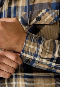 Bruine en blauwe geruite flanellen shirt met een knoopmanchet en kraag. Heeft een gestructureerde stof met contrasterende kleurpatronen en zichtbare knopen.