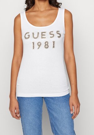 Kvinde iført en hvid tanktop med "GUESS 1981" i sølvnitter og blå jeans, stående mod en ensfarvet lys baggrund.