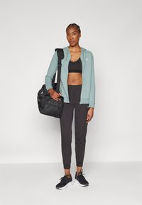 Svijetlozelena zip-up dukserica, crni sportski grudnjak, crne tapered trenirke i crne tenisice. Drži crnu sportsku torbu s mrežastim detaljima.