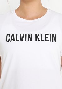 Persona vestida con una camiseta blanca con "CALVIN KLEIN" impreso en letras negras en negrita en el pecho.