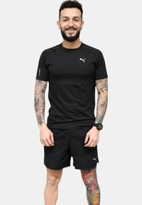 Camiseta atlética negra de manga corta con un logotipo sutil, combinada con pantalones cortos negros. El modelo muestra tatuajes y un reloj de pulsera.
