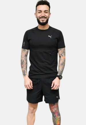 Camiseta atlética negra de manga corta con un logotipo sutil, combinada con pantalones cortos negros. El modelo muestra tatuajes y un reloj de pulsera.
