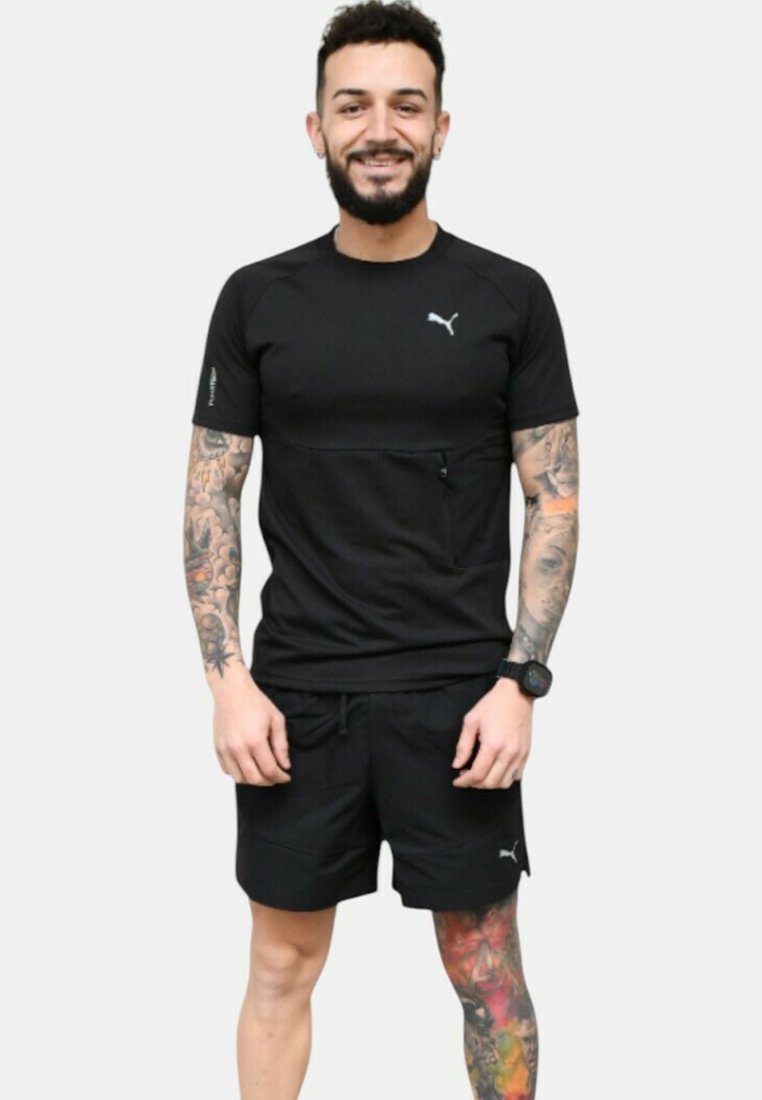 Camiseta atlética negra de manga corta con un logotipo sutil, combinada con pantalones cortos negros. El modelo muestra tatuajes y un reloj de pulsera.
