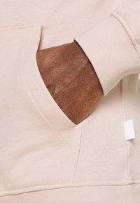 Sudadera de color rosa claro con un bolsillo tipo canguro, que muestra una tela texturizada y puños acanalados. Una pequeña etiqueta es visible cerca del dobladillo.