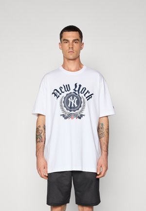 Camiseta de algodón blanca con un gráfico en azul marino que dice "New York", con un emblema y detalles florales, combinada con pantalones cortos de malla negros.