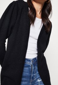 Černý pletený kardigan s texturovaným povrchem, otevřeným předním dílem a nařasenými rukávy, nošený přes bílý žebrovaný top a spárovaný s modrými džíny.