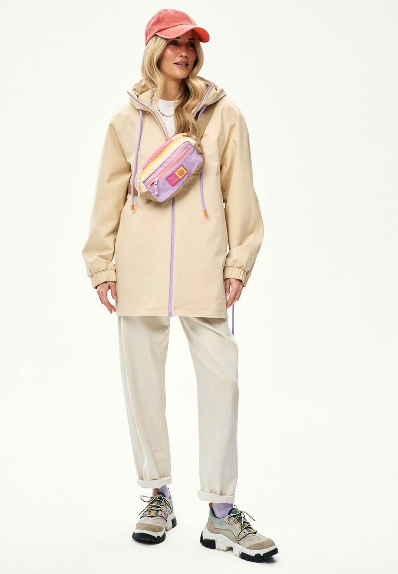 Žena oblečená v béžovej bunde s kapucňou, krémových nohaviciach, ružovej šiltovke, pastelovo farebnom ľadvinke a béžových teniskách s fialovými ponožkami, stojaca na bielom pozadí.