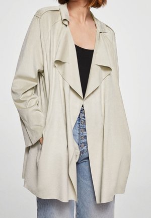 Mujer con una chaqueta suelta beige abierta por delante, sobre una camiseta negra y vaqueros de mezclilla azul claro con botones, sobre un fondo liso.