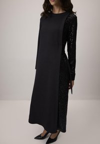 Vestido negro con un frente mate y mangas largas de lentejuelas, con un corte recto y detalle de lazo lateral. Combinado con tacones negros puntiagudos.