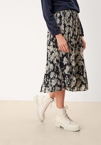 Person som bär en plisserad svart kjol med vit blommönster och vita snörstövlar som står på ett ljust beige golv.