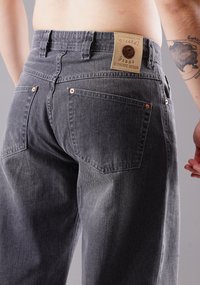 Graue Denim-Jeans mit entspannter Passform, ausgestattet mit einem beige Lederschild auf der Rückseite, Kupferriegeln und zwei rückseitigen Taschen ohne sichtbare Details.