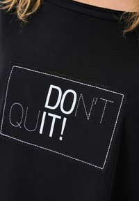 Czarny bawełniany t-shirt z prostokątną białą łatą z napisem "NIE PODDAWAJ SIĘ!" w pogrubionej czcionce, wyszytym kontrastującą białą nitką.