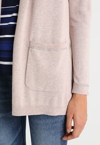 Světle růžový pletený cardigan s přední kapsou, nošený přes pruhované tričko v modrobílé barvě a modré džíny, zobrazuje pravou ruku a ruku.