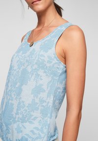 Jasnoniebieski top na ramiączkach z kwiatowym wzorem, wykonany z teksturowanej siateczkowej tkaniny, z szerokimi ramiączkami i luźnym krojem. Widoczny złoty naszyjnik z zawieszką.