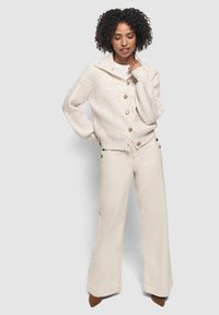Donna con i capelli ricci che indossa un cardigan a bottoni crema maculato, pantaloni larghi crema e scarpe a punta marroni, in piedi con gli occhi chiusi.