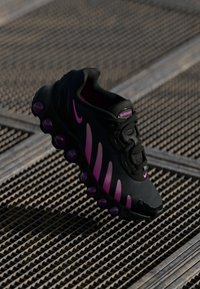 Black sneaker with pink accents, featuring a textured upper, distinctive wavy design, and rounded cushioned sole elements.