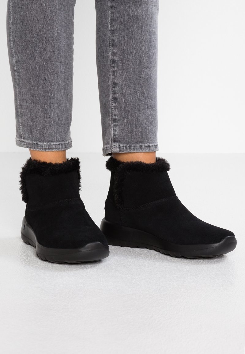 Bottines en daim noir avec une bordure en fourrure noire moelleuse, semelles en caoutchouc et un design élégant à bout arrondi, positionnées sur un fond blanc.
