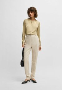 Lichtgroene blouse met lange mouwen en geplooide halslijn, gecombineerd met beige hoge taille broeken. Zwarte puntige hakken en een kleine handtas.