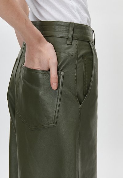 Χέρι που έχει εισαχθεί στην πίσω τσέπη σκούρων πράσινων δερμάτινων παντελονιών, φορεμένων με λευκό ριμπ τοπ, σε απλό φόντο.