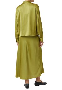 Person wearing a lime green satin long-sleeve blouse and matching long skirt with black studded Mary Jane shoes, viewed from behind.