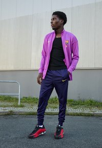 Paarse Nike-jack met het Barcelona-logo, zwarte T-shirt, marineblauwe broek met gele accenten, en zwarte/rode Nike-schoenen. Staand buiten op het asfalt.