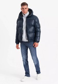 Giacca a vento blu navy con cappuccio, design trapuntato, zip frontale, polsini elasticizzati e toppa con logo. Indossata sopra una felpa grigia chiara e jeans blu.