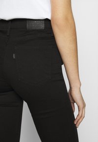 Jeans de mezclilla negra con corte slim, bolsillos traseros con costura curva y un parche de cuero en la cintura. Textura suave y líneas limpias.