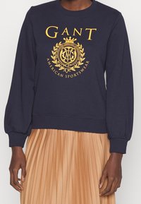 Felpa blu navy con scollatura rotonda e maniche a sbuffo, caratterizzata da un grande logo giallo e un emblema centrati sul petto. Polsini aderenti.