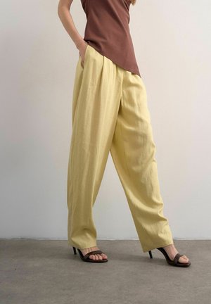 Kvinde iført løstsiddende gule bukser med høj talje, brun ærmeløs top og brune åben tå hælede sandaler, stående mod en almindelig væg.