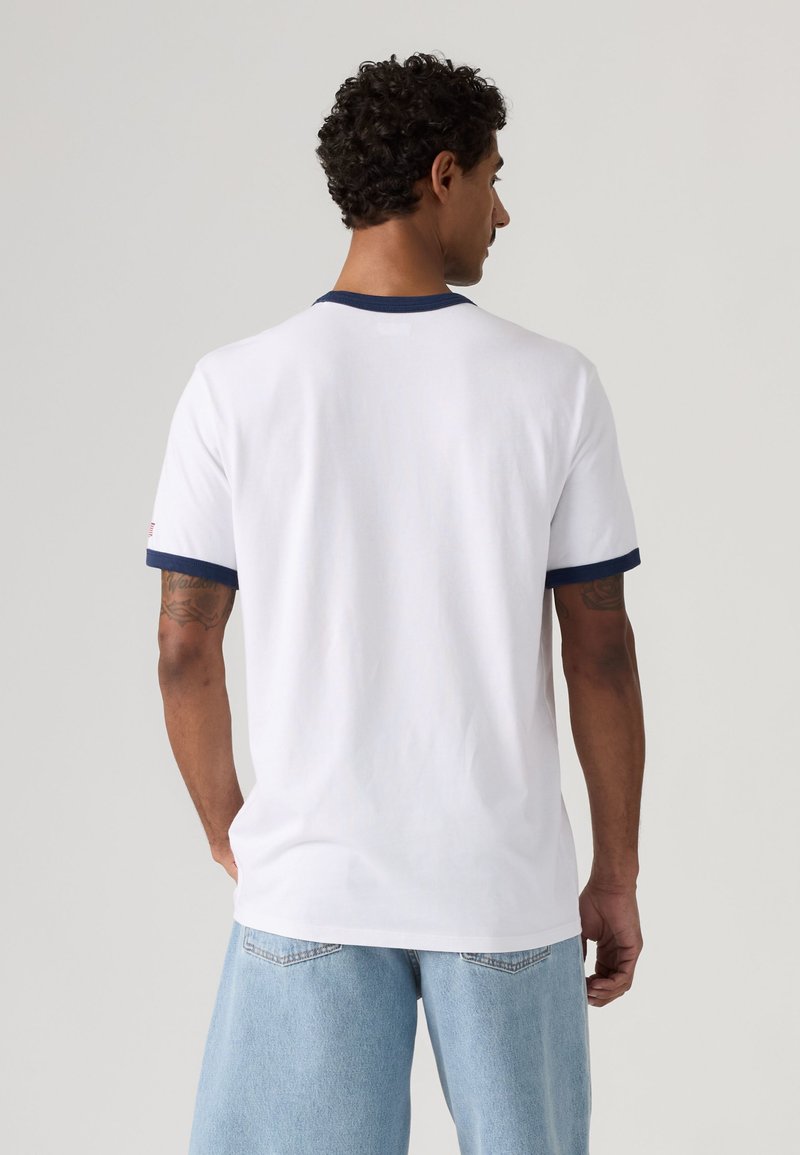 Man with curly hair wearing a white t-shirt with navy trim and light blue jeans, standing facing away against a plain background.