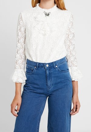 Kvinde iført hvid blondebluse med flæsekrave og butterflybroche, kombineret med højtaljede blå jeans mod en ensfarvet baggrund.