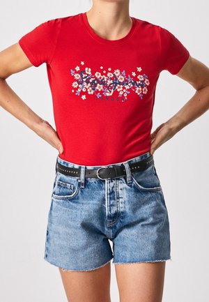 Osoba nosząca czerwony t-shirt z kwiatowym nadrukiem włożony do niebieskich, wysokiego stanu szortów jeansowych z czarnym paskiem, ręce na biodrach.