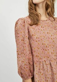 Femme portant une robe rose avec un petit imprimé floral jaune et rouge, manches bouffantes et poignets froncés.
