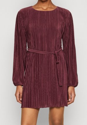 Kvinde iført en langærmet, plisseret minikjole i mørk mauve med et matchende stofbælte bundet i taljen.