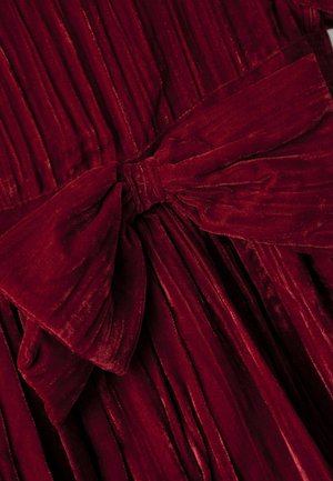 Tumši sarkans samta audums ar lielu, teksturētu loku, kas sasiets pār krokotā materiāla, izceļot bagātīgu tekstūru un krokas.