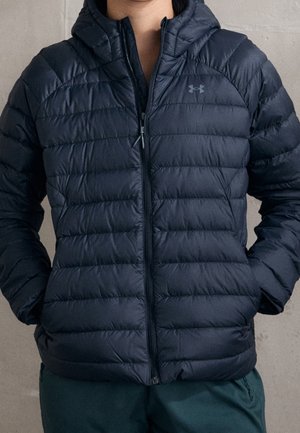 Person trägt eine dunkelblaue gesteppte Under Armour Jacke mit Frontreißverschluss und die Hände in den Taschen vor neutralem Hintergrund.