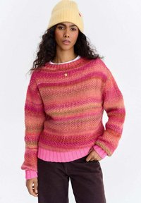 Kobieta w żółtej dzianinowej czapce, różowo-pomarańczowym swetrze w paski, ciemnych spodniach i złotym naszyjniku, stojąca z jedną ręką w kieszeni.