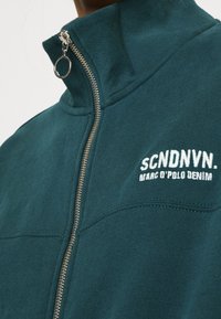 Teal-Fleecejacke mit Reißverschluss, hochgeschnallt und silbernem Reißverschluss. Auf der linken Brust ist das Logo in weißem Faden gestickt.