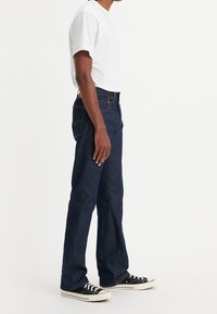 Jean en denim bleu foncé avec des coutures visibles, une coupe droite et un design classique à cinq poches. Portés avec un t-shirt blanc et des baskets noires.