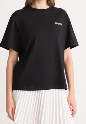 Femme portant un T-shirt noir à manches courtes avec un petit logo vert sur la poitrine, associé à une jupe plissée blanche sur fond uni.