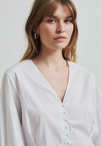 Blusa bianca realizzata in tessuto morbido, con scollatura a V, maniche lunghe e una chiusura con bottoni al centro con piccoli bottoni coordinati.