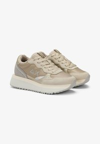 Beige lace-up sneakers with thick white sole, soft textured lining, stitched geometric logo on side, and ridged outsole for grip.