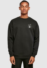 Must rõngas krae ja pikkade varrukatega musta pehmest kangast sweatshirt. Vasakul rinnal on väike valge graafika.