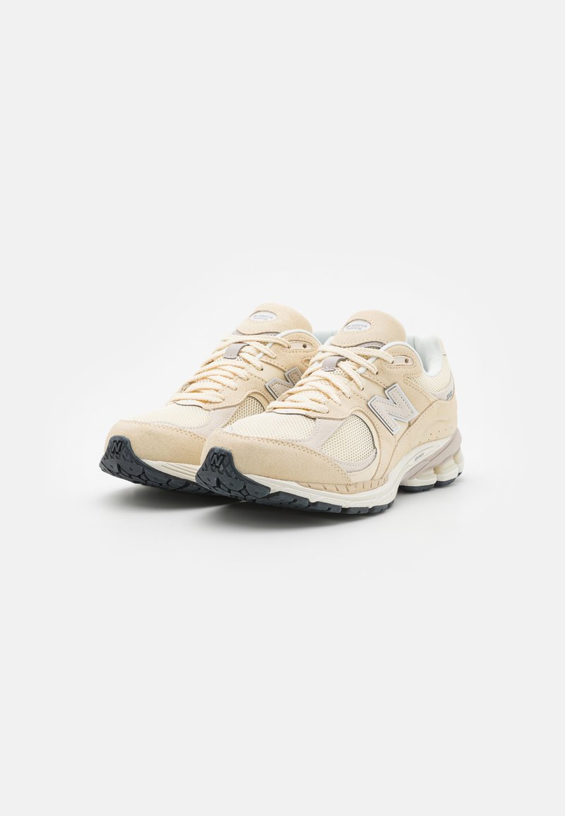 Un paio di scarpe da corsa New Balance beige e crema con lacci beige e suole nere su uno sfondo bianco.