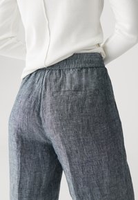 Personne portant un pantalon bleu texturé avec une taille élastique et un haut à manches longues côtelé blanc, les mains posées sur les hanches.