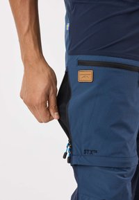 Marinblå utomhusbyxor med en blixtlåsförsedd ficka, med en tanfärgad läderlapp märkt "SWEDMOUNT" och en kontrasterande blå dragkedja.