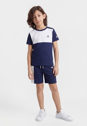 Junge mit mittellangen Haaren steht vor weißem Hintergrund und trägt ein marineblau-weißes kurzärmeliges Hemd, passende Shorts und weiße Turnschuhe.