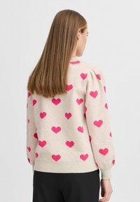 Pull beige clair avec un motif répété de cœurs rose vif. Des manches bouffantes et un ourlet côtelé complètent le design.